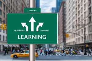 Grünes Schild mit Aufschrift "LEARNING" und drei Pfeilen in verschiedene Richtungen. Im Hintergrund eine New-Yorker Straßenszene.